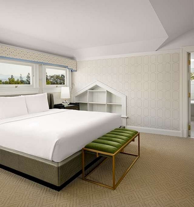 A bright hotel room with a king bed, matching nightstands, a bench at the foot, windows, and a small shelf nook by the wall.