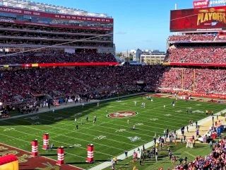 A packed football stadium with red-clad fans, a bright green field, and clear blue sky during a game.