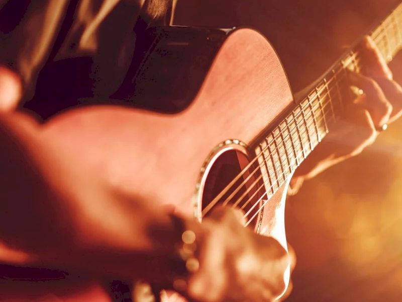 A close-up of a person playing an acoustic guitar with warm stage lighting, focusing on the musician&rsquo;s hands and strings.