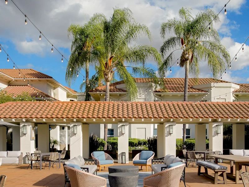 A sunny courtyard with palm trees, string lights, and outdoor seating around a fire pit in a tiled patio.