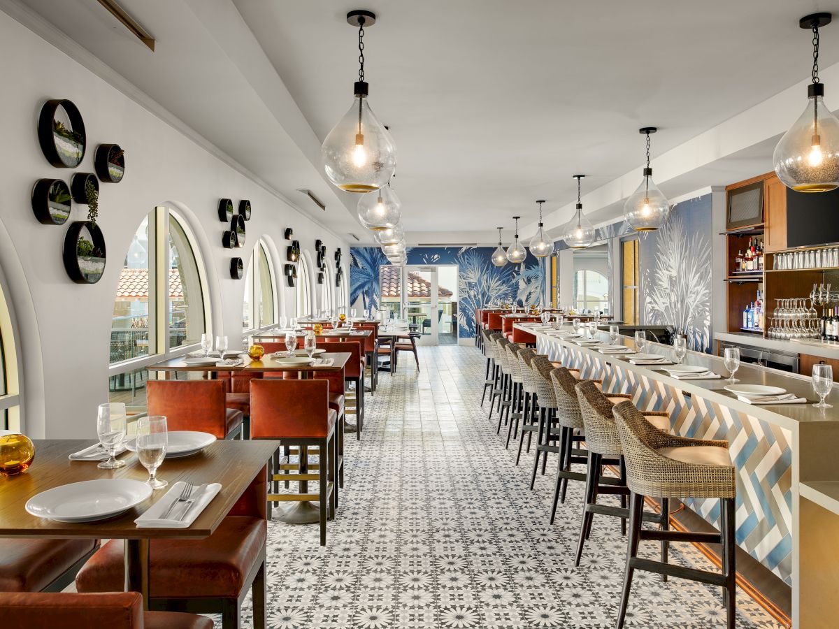 A stylish, airy restaurant with long wooden tables, vintage-style chairs, geometric tiled floor, and warm pendant lighting, ready for diners.