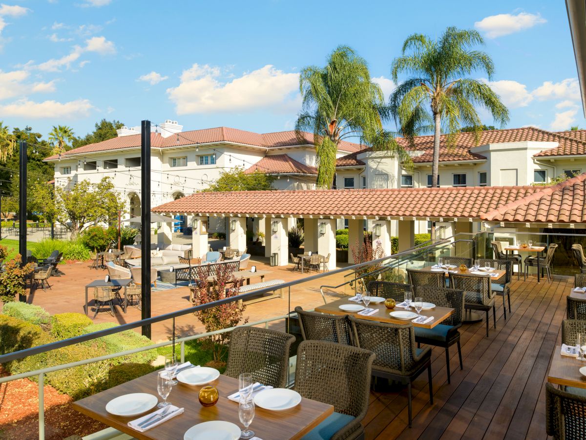 An outdoor resort terrace with wooden decking, dining tables set for meals, palm trees, sunlit buildings, and a clear blue sky.