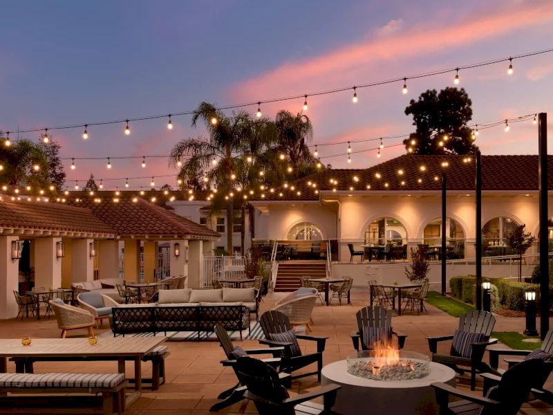 A cozy outdoor courtyard with string lights, lounge chairs, a fire pit, palms, and warm sunset skies.