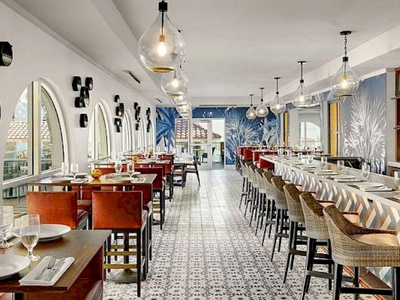 A bright, stylish dining area with long tables, pendant lights, arched windows, and patterned tile floor, ready for a crowd.