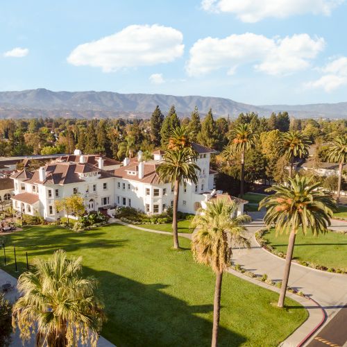 A sunny campus or resort with well-kept lawns, palm trees, white buildings, and a winding driveway under a blue sky with fluffy clouds.
