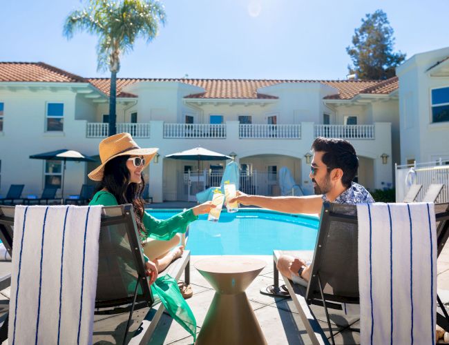 Two people toast by a pool at a sunny resort, lounging on striped chairs with a palm tree and a white building in the background.