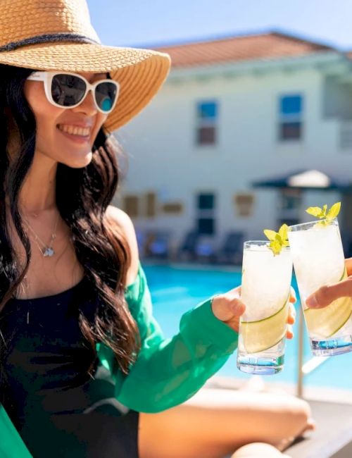 Two women clink tropical cocktails by a pool, one wearing a sunhat and sunglasses, smiling under sunny resort vibes.