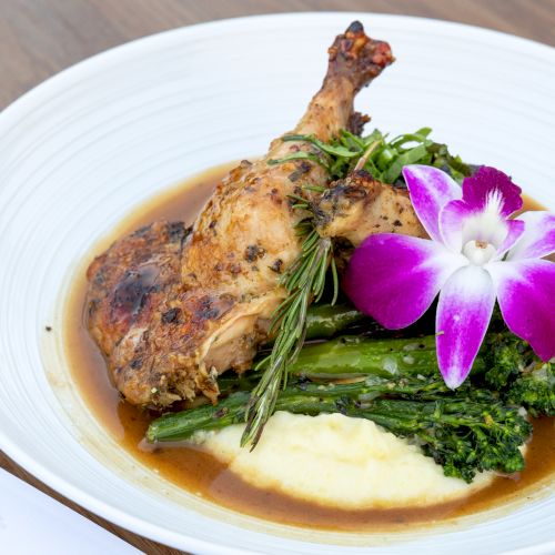Roasted chicken leg on greens with sauce, mashed potatoes, and a purple orchid garnish on a white plate.