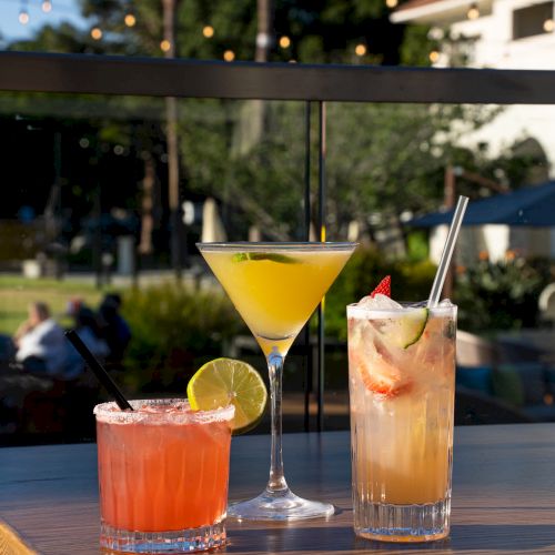 Three colorful cocktails on a patio table with lime garnish and iced drinks, sunset lighting, and a blurred outdoor scene in the background.