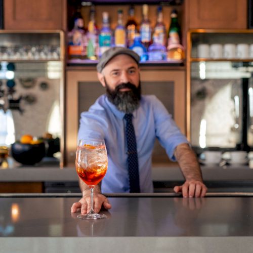 A bartender with a beard and cap stands behind a bar, pointing a glass of orange drink toward the front, inviting a toast.