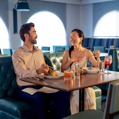 Two adults sit at a booth in a stylish restaurant, chatting and laughing with drinks and plates in front of them.