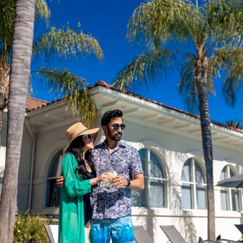 A couple stands by a white villa with palm trees, sunny sky, and poolside chairs; the man wears a floral shirt, the woman in a green dress and hat, both smiling.
