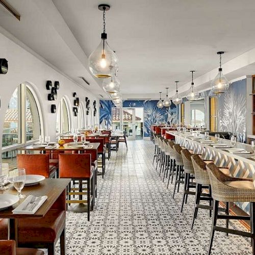 A stylish, bright restaurant with a long bar on the right, arched mirrors on the left, warm wood tables, and geometric tiled floor, modern pendant lights.