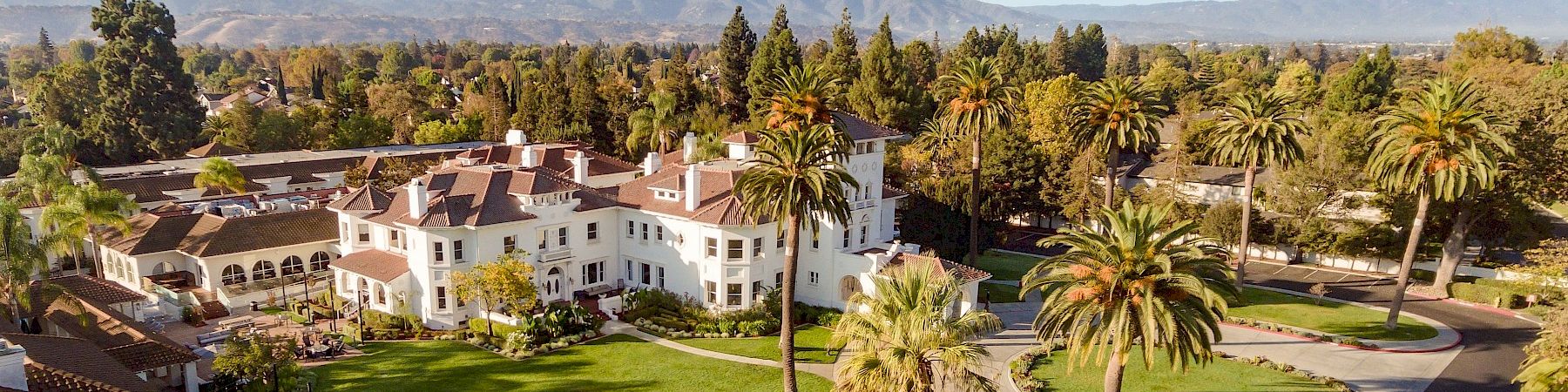 A sunny resort-style campus with white buildings, palm trees, green lawns, and winding paths under a blue sky with scattered clouds.