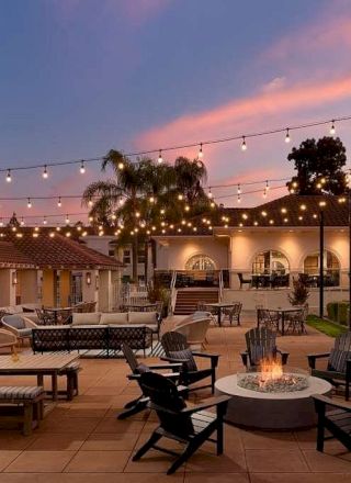 Outdoor hotel courtyard at dusk with string lights, lounge chairs, benches, and a circular fire pit surrounded by palm trees and buildings.