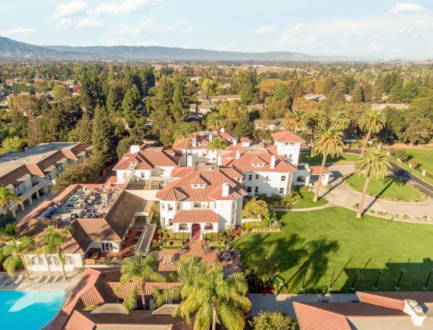 Aerial view of a resort with a large pool, sun loungers, palm trees, and a cluster of colorful buildings amid green lawns and clear skies.
