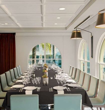 A bright, elegant dining room with a long glass table, stylish chairs, a chandelier, and large windows letting in natural light.